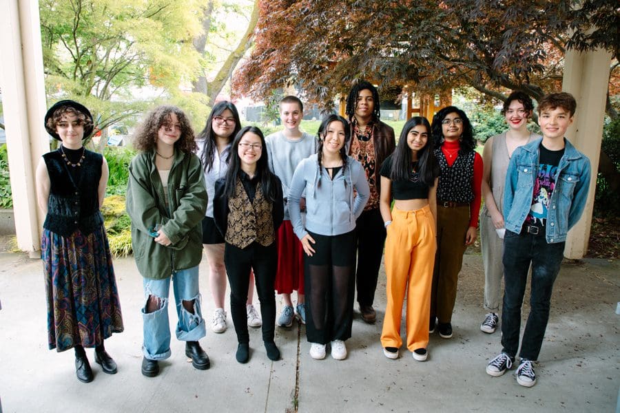 The current Youth Poet Laureate cohort, a diverse group of people aged 19 and younger, stand in a circle with their hands piled together.