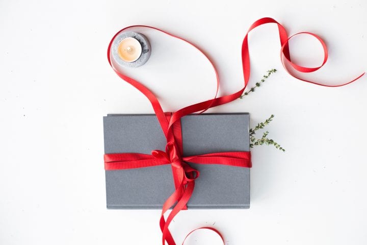 A grey book is resting against a white background, with green sprigs tucked into its pages. It's wrapped in a bright red curling ribbon.