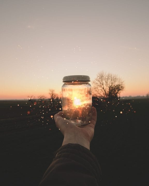 Hand holds a mason jar with a lid on it up to the setting sun